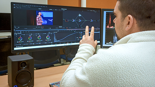 Im Studium wie die Profis arbeiten: Postproduction mit DaVinci Resolve Studio Person an einem Schreibtisch von hinten im Anschnitt vor einem großen Monitor.